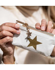 A close-up of a person holding a small white zip pouch with a large metallic gold star on the front and a matching gold zipper pull charm.