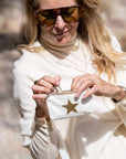 A woman wearing a cream turtleneck sweater and tortoiseshell sunglasses holds a small white pouch with a gold star in a sunny outdoor setting.