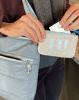 A close-up lifestyle shot of a person holding a light gray quilted shoulder bag and reaching into a small beige coin pouch with a silver 'H!' logo. The person is wearing a brown sweater and dark pants, and the scene is set in an indoor environment with a marble countertop in the background.