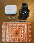 Orange Bandana pattern clutch with black watch, ear buds, and earrings on a wooden surface