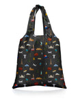 A reusable folding tote bag with a black background and an all-over travel-themed print featuring landmarks from the seven continents along with airplanes, a globe, mountains, a passport and a luggage in small multicolor icons, shown on a clean white background.