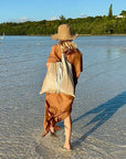Person walking along a beach with a beige tote bag, straw hat and brown dress, surrounded by greenery and water.