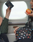 Two women smiling on a plane, holding a black travel-themed lunch bag with a colorful world landmark print. One woman holds a passport, and the other holds an apple.