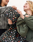 Two women smiling and holding a black reusable tote bag and lunch box with a colorful travel-themed pattern, set against a bright, overexposed outdoor background.
