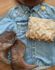 Medium Shimmer Bronze Bandana Print zipper pouch styled on a denim shirt beside brown cowboy boots and brown hat.