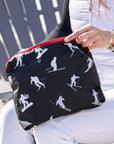 A black zippered pouch with a white ski and snowboard silhouette pattern and a red zipper, held by a person wearing white ski pants and a white top in an outdoor setting.
