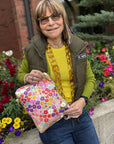 Woman holding a colorful, floral-patterned zipper pouch outdoors with flowers and a building in the background