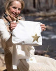 A woman outdoors by a flowing river holds up a white bag or pouch with a large metallic gold star on the front. She is wearing a cream sweater and a patterned scarf, and the background shows blurred rocks and water.