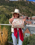 Women wearing a western hat and red silk scarf in the mountains holding a zipper pouch with western themed accents printed on it.