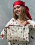 Young woman standing against a white wall wearing a red silk scarf holding a western themed pouch with cowboy boots, cactus, horses, and ropes printed on the pouch.