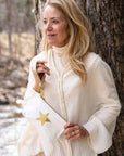 A woman standing outdoors next to a tree, wearing a cream ribbed turtleneck sweater and holding a white zippered pouch bag with a gold star detail.
