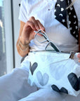 A person holding a white zippered pouch with a black and gray heart pattern, holding a pair of eyeglasses inside. The person is wearing a white t-shirt and white pants.