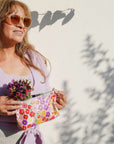 Woman holding a colorful floral clutch and flowers against a white wall with shadows.