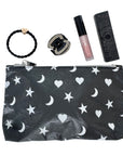 A black cosmetic pouch with a white celestial pattern of stars, hearts, and crescent moons, displayed alongside beauty accessories including a black braided hair tie, a rhinestone ring, a lip gloss tube, and perfume on a white background.