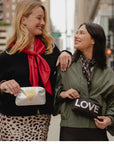 Two women standing outdoors on a city street, each holding a black clutch bag with Gold Heart and LOVE text. The woman on the left wears a black top and a bright red scarf, and the woman on the right wears an olive green jacket and a dark patterned scarf.