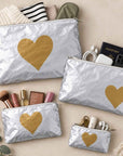 Set of four silver travel pouches with gold hearts on beige background holding various beauty and travel itrems