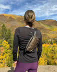 Person standing wearing Puffer Crossbody Fanny Pack Belt Bag in Shimmer Bronze with fall mountain background