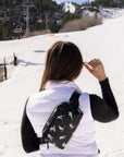 A woman on a ski slope seen from behind, wearing a black crossbody belt bag with a white ski pattern over a white puffer vest and black long-sleeve shirt.