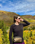 Woman smiling wearing Puffer Crossbody Fanny Pack Belt Bag in Shimmer Bronze with fall mountain background