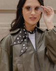 A woman wearing a black neck scarf with a white crescent moon, heart and star pattern, styled with an olive green leather jacket and clear-framed glasses.