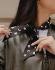 A close-up of a woman wearing a black satin scarf with a white celestial pattern of stars, hearts and crescent moons, styled under a dark olive green leather jacket.