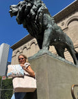 A woman stands outdoors beside a large bronze lion statue on a stone pedestal, holding a large beige tote bag and pouch with colorful illustrated Chicago landmarks. A beige building and blue sky are visible in the background.
