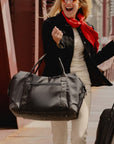 A woman in a black blazer and cream pants carries a large black duffel bag on a city walkway, with a bright red scarf and part of another person's arm visible.