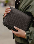 A close-up of a person holding a shimmer black textured clutch bag with a repeating embossed monogram pattern. The person is wearing an olive green jacket and a gold link bracelet, with a blurred outdoor background.