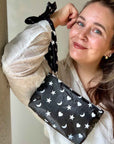 Woman wearing a black silk scarfette as a wristlet strap attached to a mini zipper pouch with white hearts, moons and stars pattern.