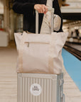 Woman traveling with a beige Signature travel tote on a silver rolling suitcase at a train platform.
