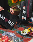 Woman holding black with "AMORE" zipper pouch surrounded by picnic foods