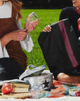 Two women on picnic blanket holding black wine tote and glass water bottle