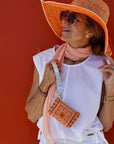 Woman wearing a large orange hat, sunglasses, and holding a orange bandana pouch against a red background