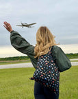 Person standing in field waving at airplane in the sky holding black tote bag with travel inspired pattern of landmarks from the seven continents.