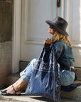 Woman sitting on doorstep wearing denim outfit and black hat holding navy bandana print tote bag