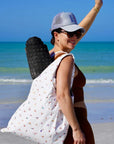 Woman on a beach with a white bag with yoga characters and gray cap, smiling.