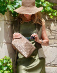 Woman wearing hat and army green dress pulling sunglasses out of woven shimmer bronze clutch