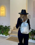 Person wearing a black hat and silver backpack walking into a building with plants and signage.