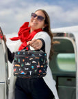 Woman standing on plane in red scarf and sunglasses, smiling holding black travel inspired lunch box.
