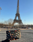Hand holding a travel patterned pouch in front of the Eiffel Tower