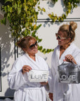 Two women in white robes holding 'LOVE' bags outdoors.