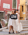 Two women standing in front of colorful buildings in Italy holding large black with silver love zipper pouch