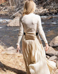 A woman stands outdoors by a flowing river, shown from the back. She is wearing a fitted beige mock-neck long-sleeve top, a matching pleated maxi skirt, and a shiny metallic gold belt bag with a black strap.