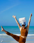 Person in brown outfit with a hat and pouch on their head, standing on one leg on a beach with blue sky and ocean.