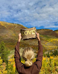 Person standing in a valley with foliage color changes holding bronze pouch with gold LOVE
