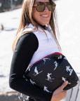 Woman wearing white vest and a black top in an outdoor setting, holding black zippered pouch with a white ski and snowboard silhouette pattern and a red zipper.