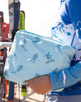 Close-up of a light blue medium zipper pouch featuring an all-over skier print, held by a woman in ski apparel at a mountain resort.