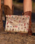 A beige pouch with western boots, hats, horse and cactus on it in an outdoor setting with brown cowboy boots worn by two people.