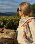 A woman in profile wearing sunglasses, a cream cable-knit sweater, and a colorful patterned scarf, holding a metallic bronze clutch with gold moon and star designs against a scenic outdoor background of mountains and greenery.