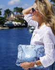 Woman holding a blue bandana clutch by a waterfront with palm trees in the background